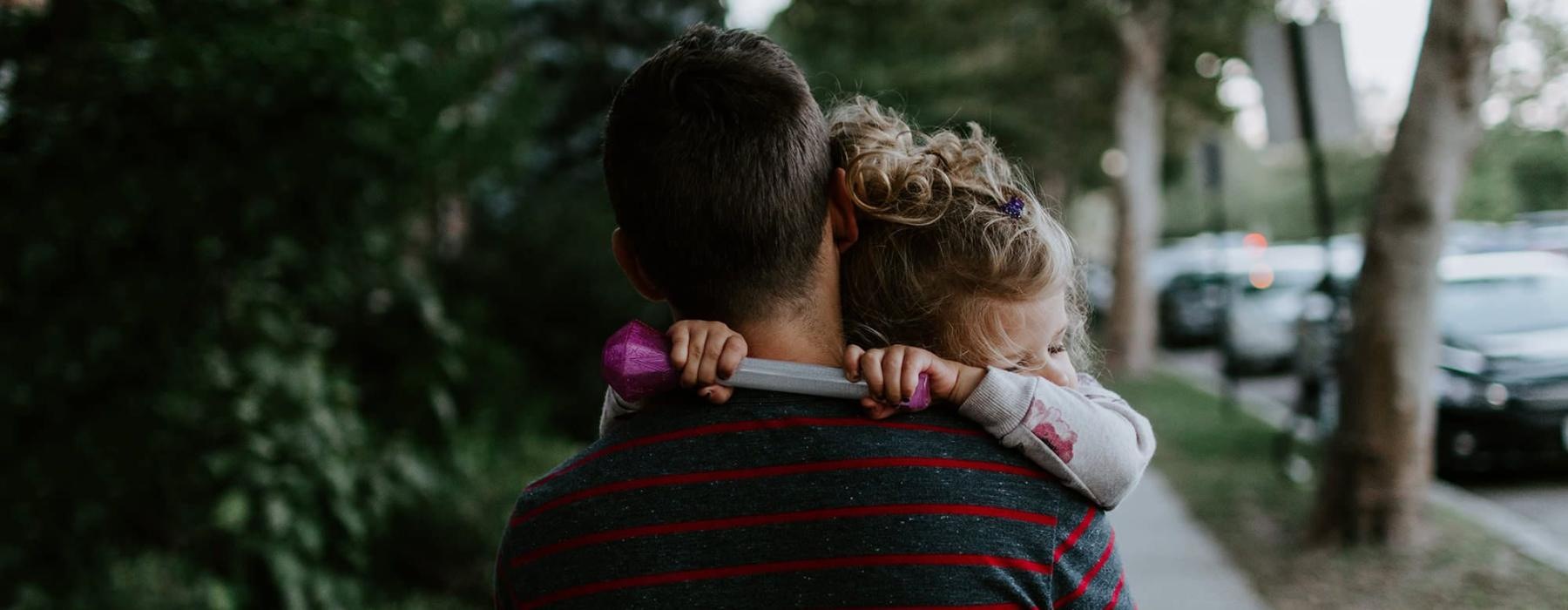 tired little girl rests her head on her father's shoulder as he carries her down the city sidewalk