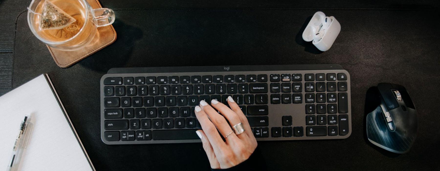 woman's hand on a keyboard surrounded by office items and a cup of tea on a coaster